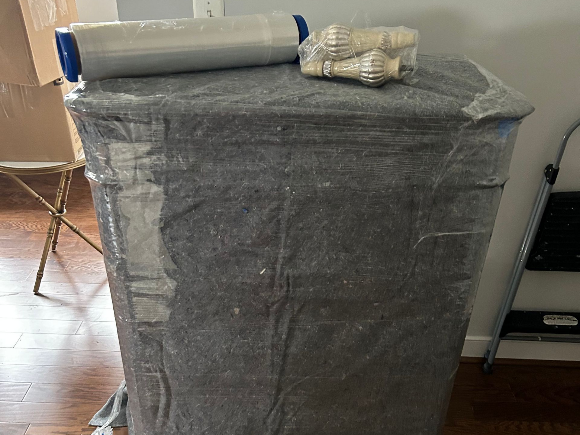 Wrapped table in gray material with plastic wrap, tape, and packing supplies on top.