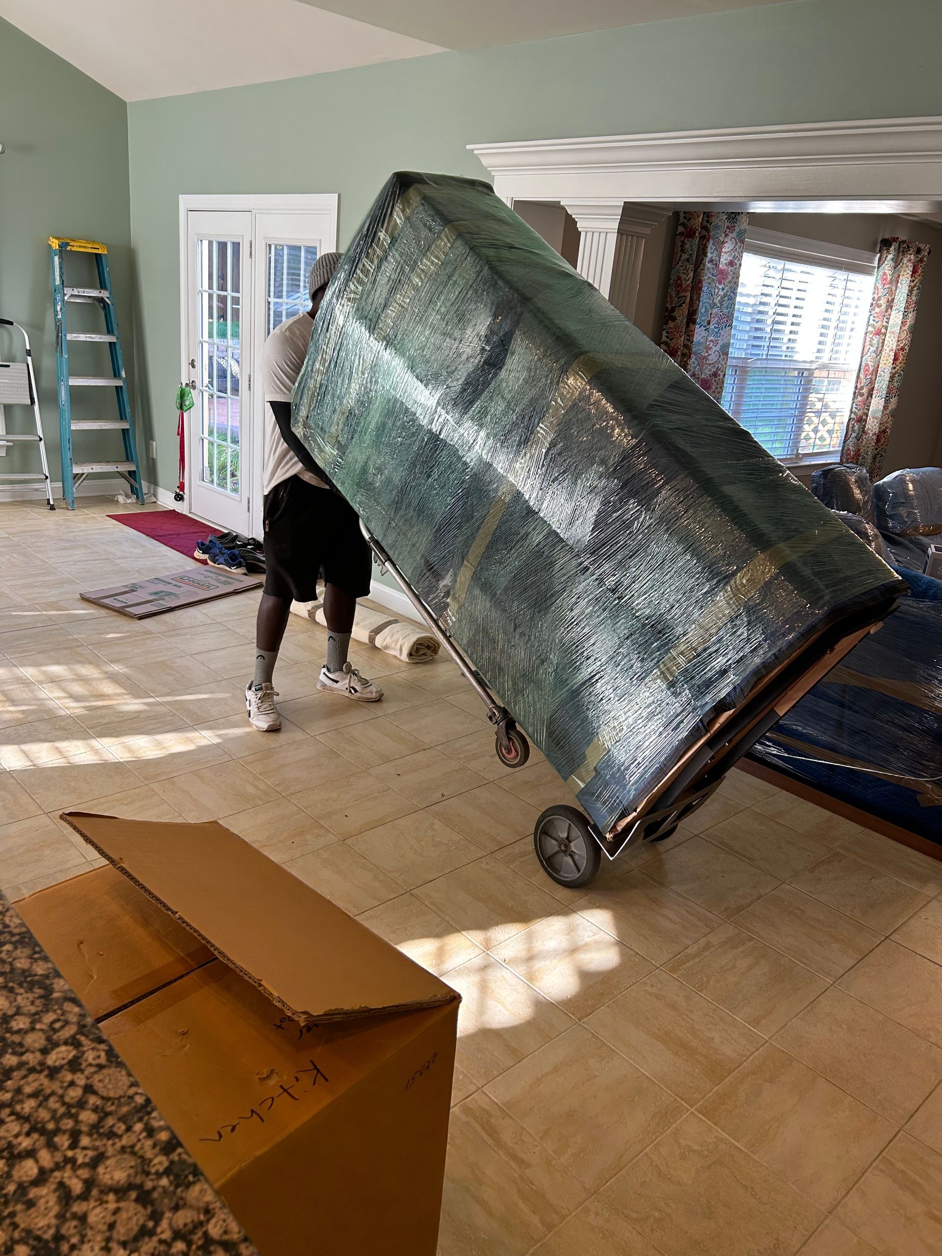 Person using a dolly to move a large, wrapped piece of furniture indoors. Boxes are on the floor.
