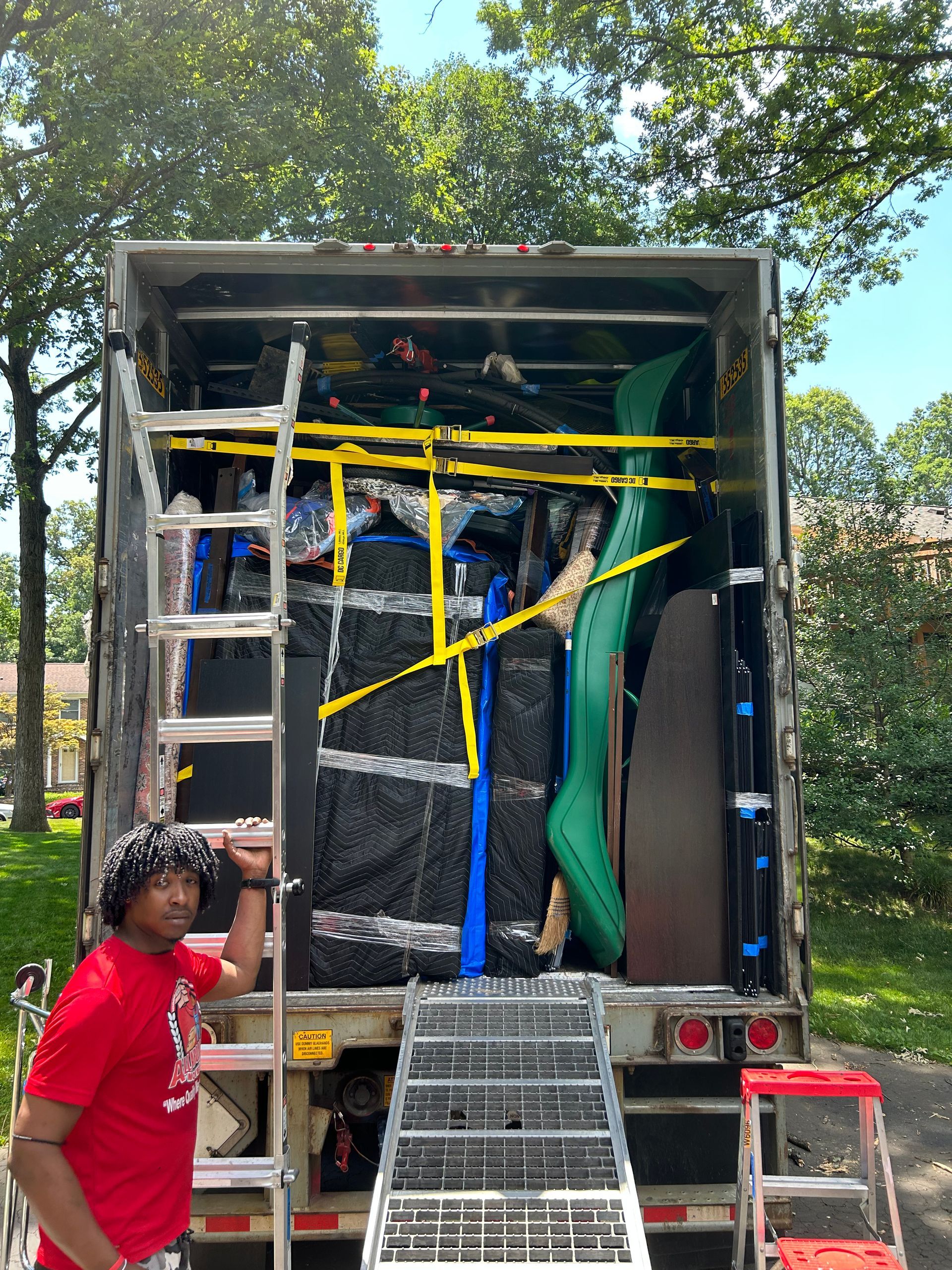 A person in a red shirt stands next to a moving truck packed with furniture and a bright green kayak, under green trees.