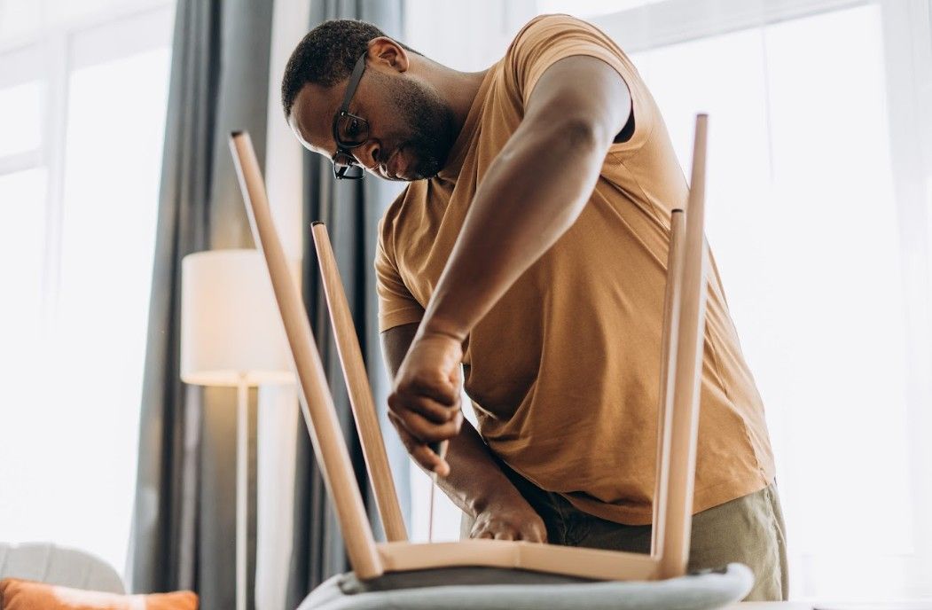 Man assembling a chair with a screwdriver, indoors, near a window.