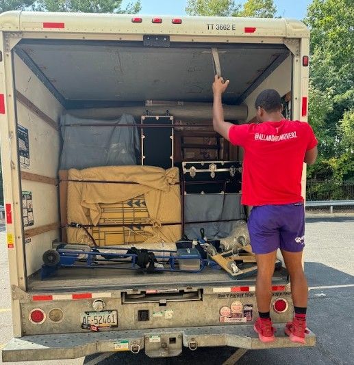 Man in red shirt inside the back of a moving truck with cargo, holding a tool up.