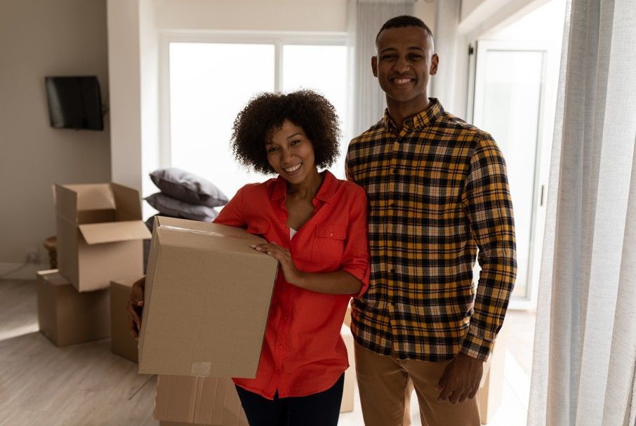 Smiling couple holding a cardboard box in a room with packed moving boxes.