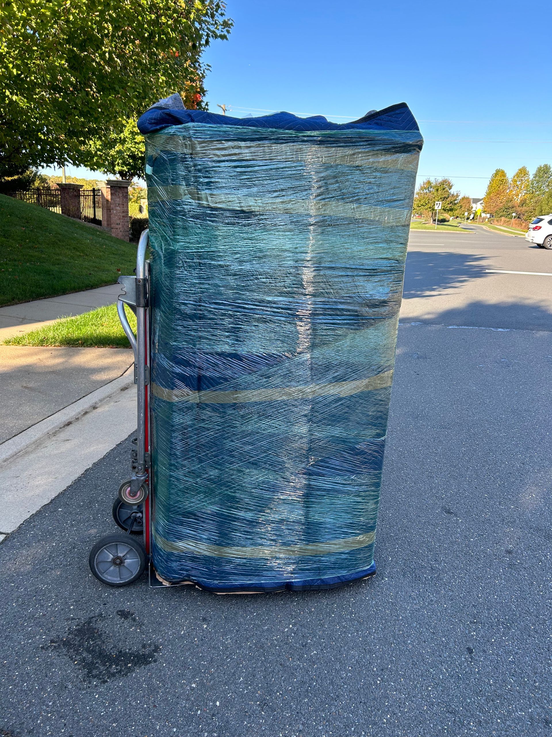 A large piece of furniture wrapped in plastic is on a dolly on the side of the road.