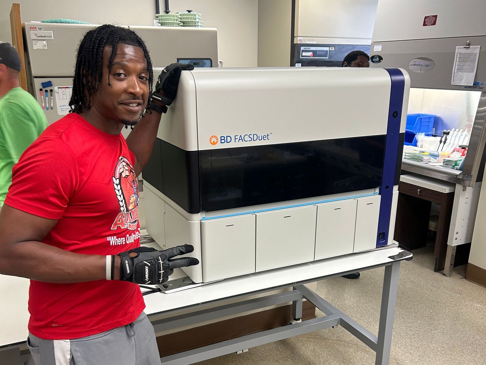 A man in a red shirt is holding a machine in a lab.