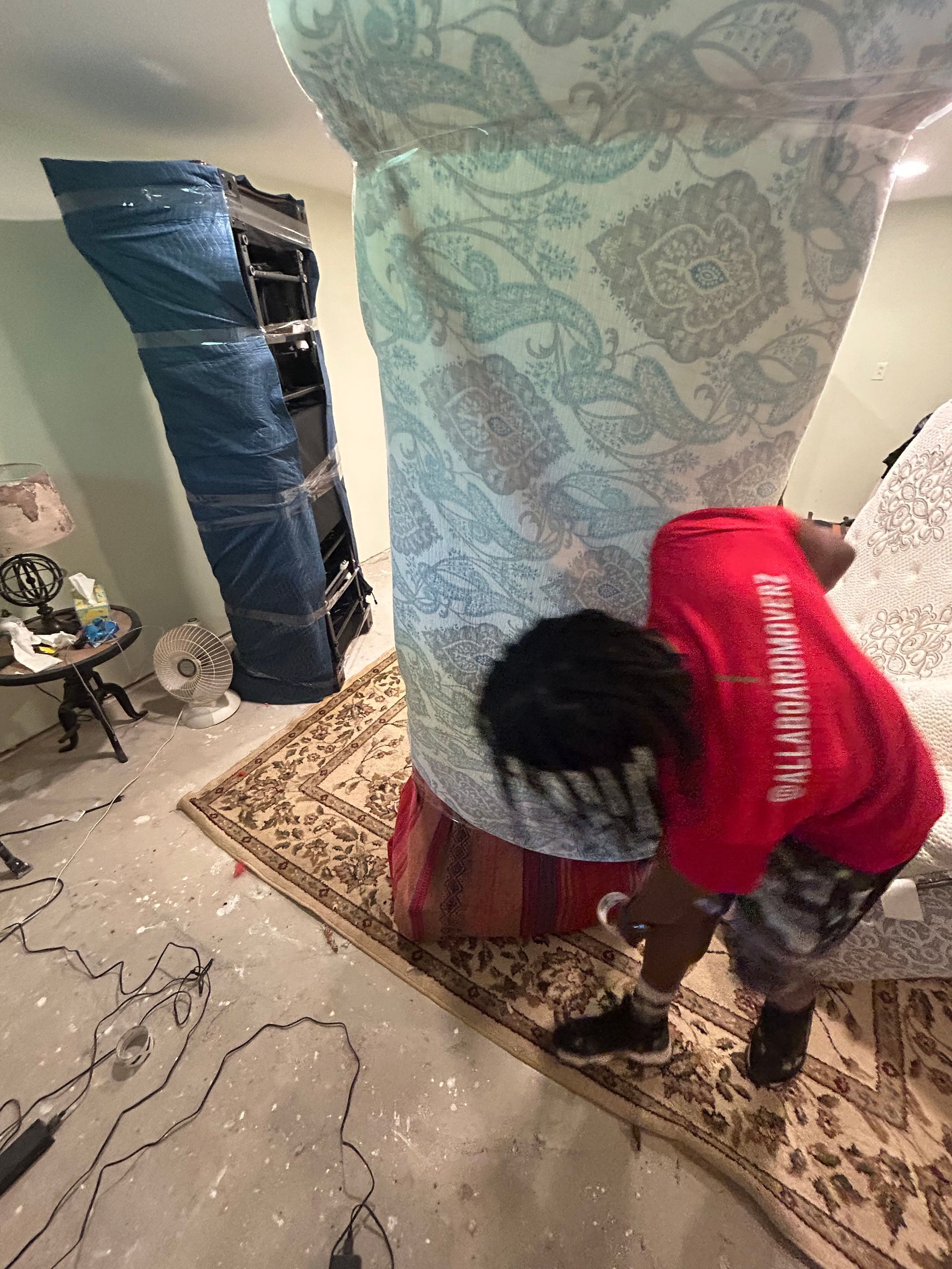 A man in a red shirt is standing next to a large mattress in a room.