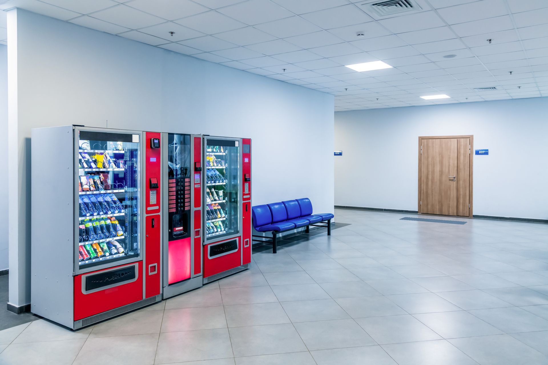 Vending machines and blue bench in a well-lit hallway with a wooden door.