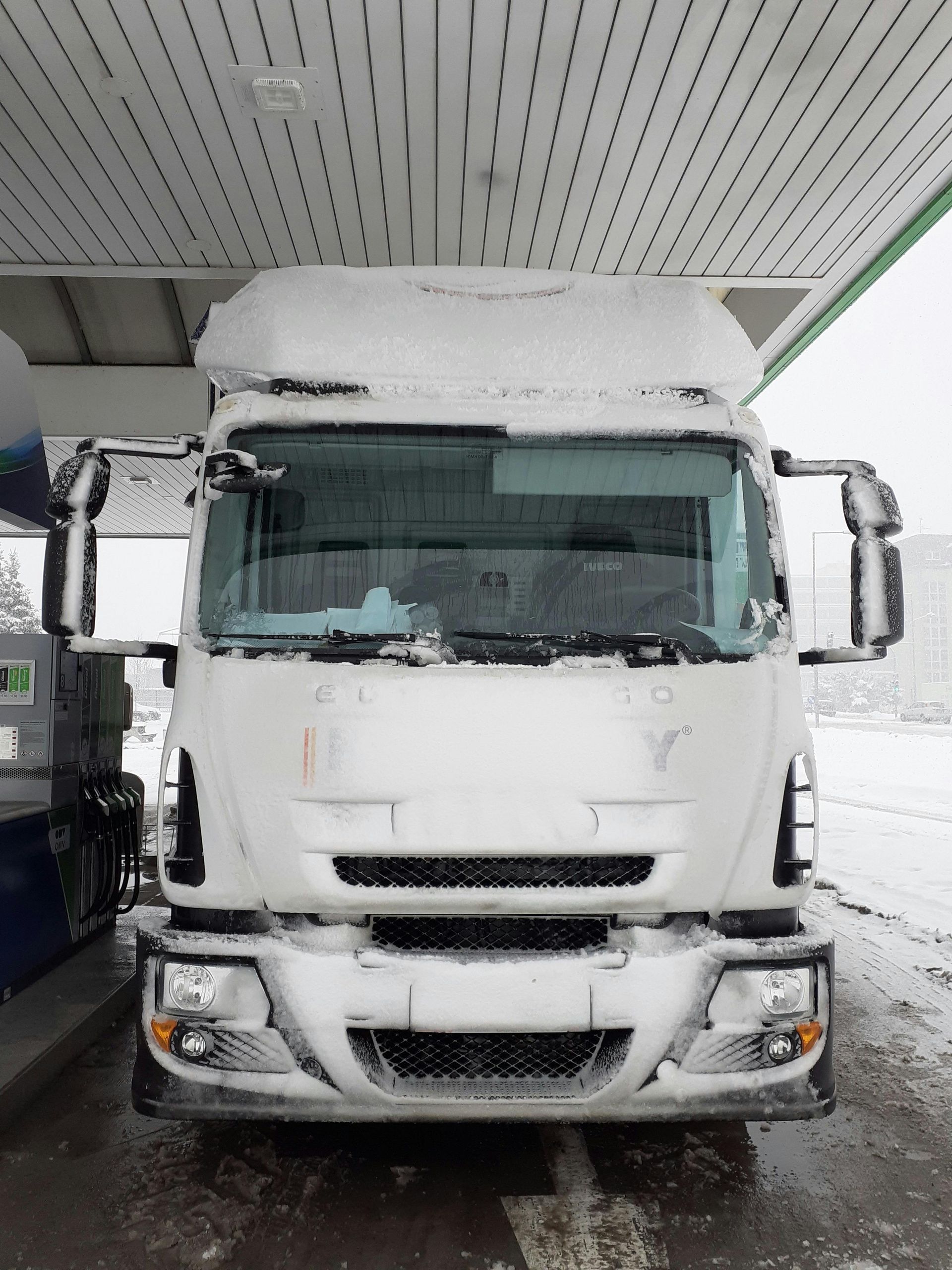 Snow-covered white semi-truck parked under a gas station canopy.