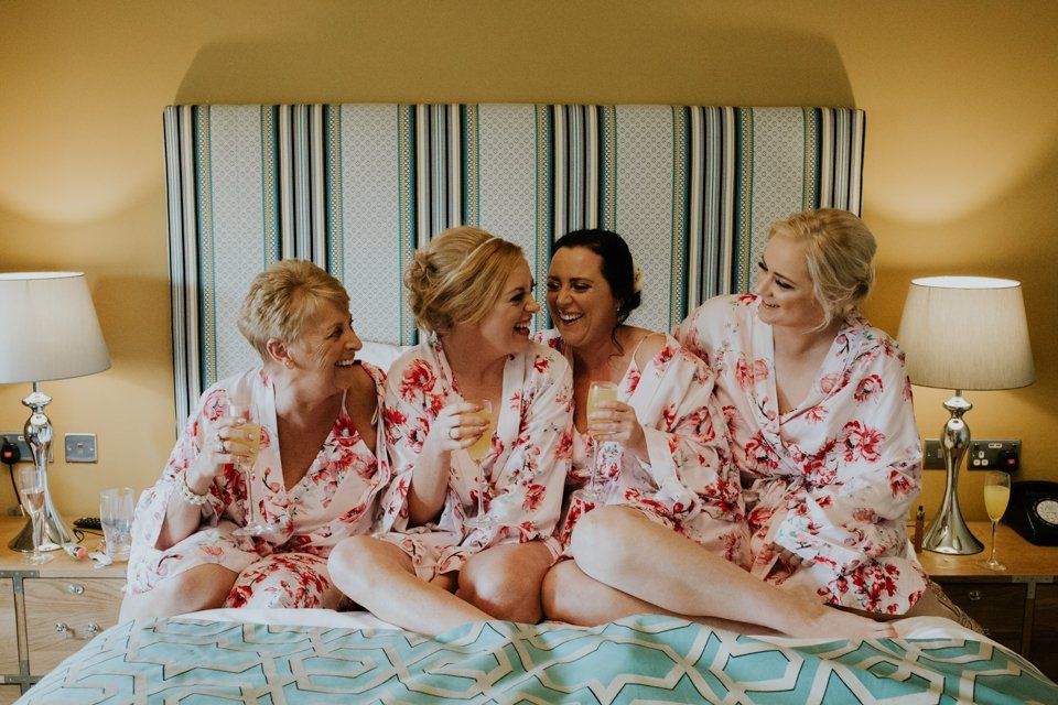 bridesmaids sat together on hotel bed drinking champagne and laughing together before wedding