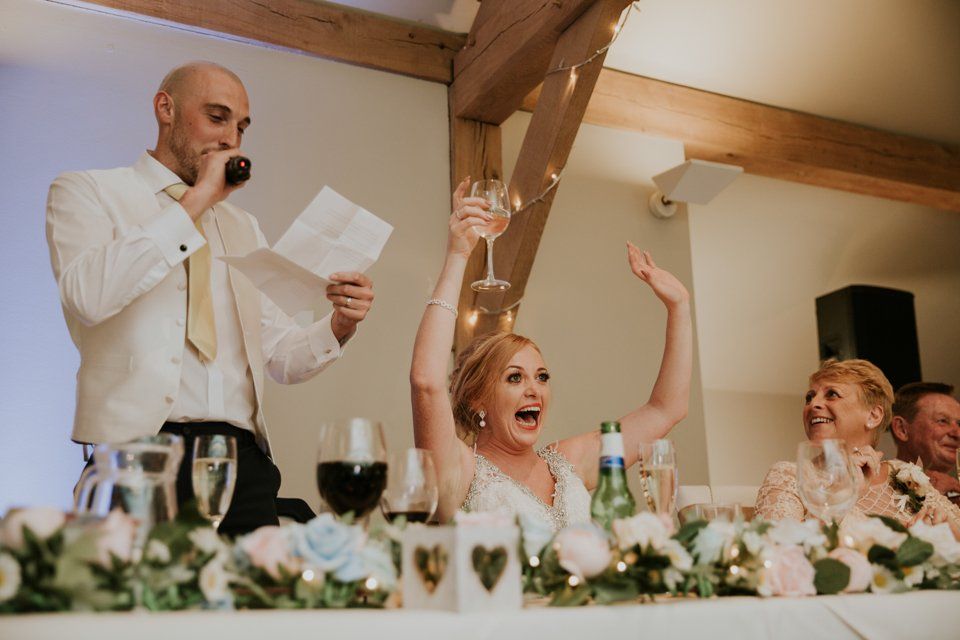 bride smiling with her hands in the hair whilst the groom reads his speech
