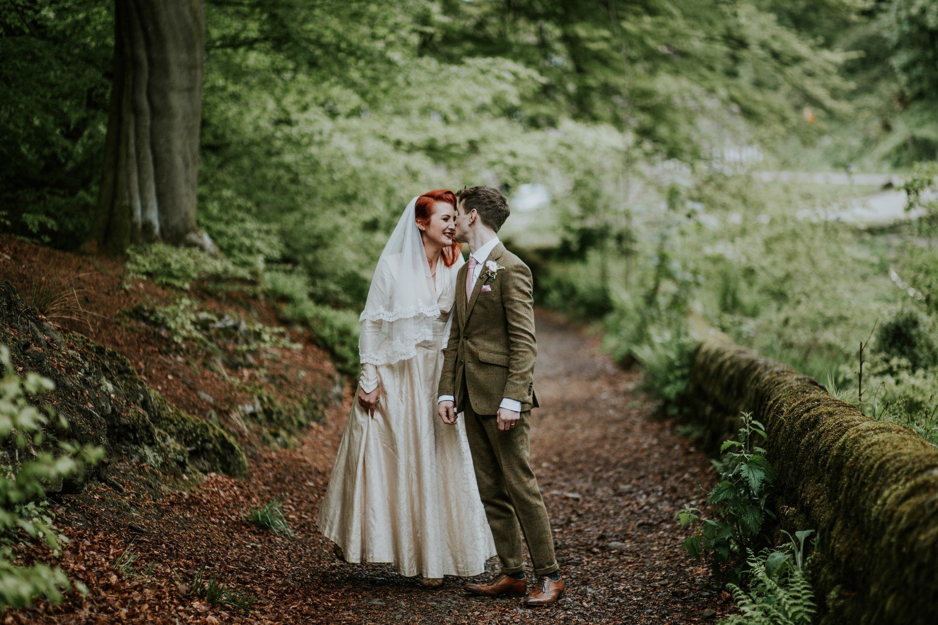 wedding photograph of bride and groom holding hands and kissing on path in a forest