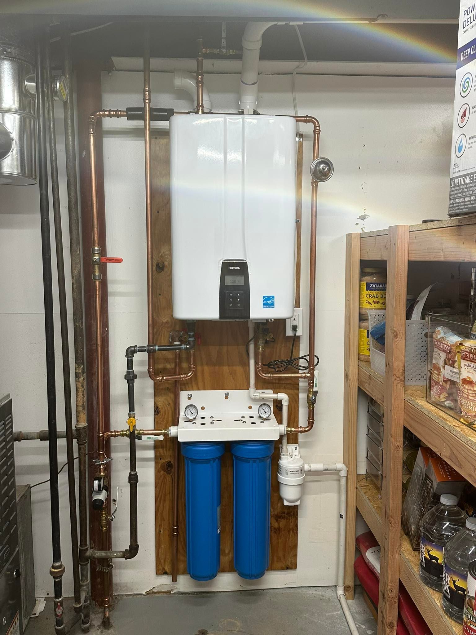 Tankless water heater and water filter system mounted on a wood panel in a garage. Copper pipes and blue filter housings.