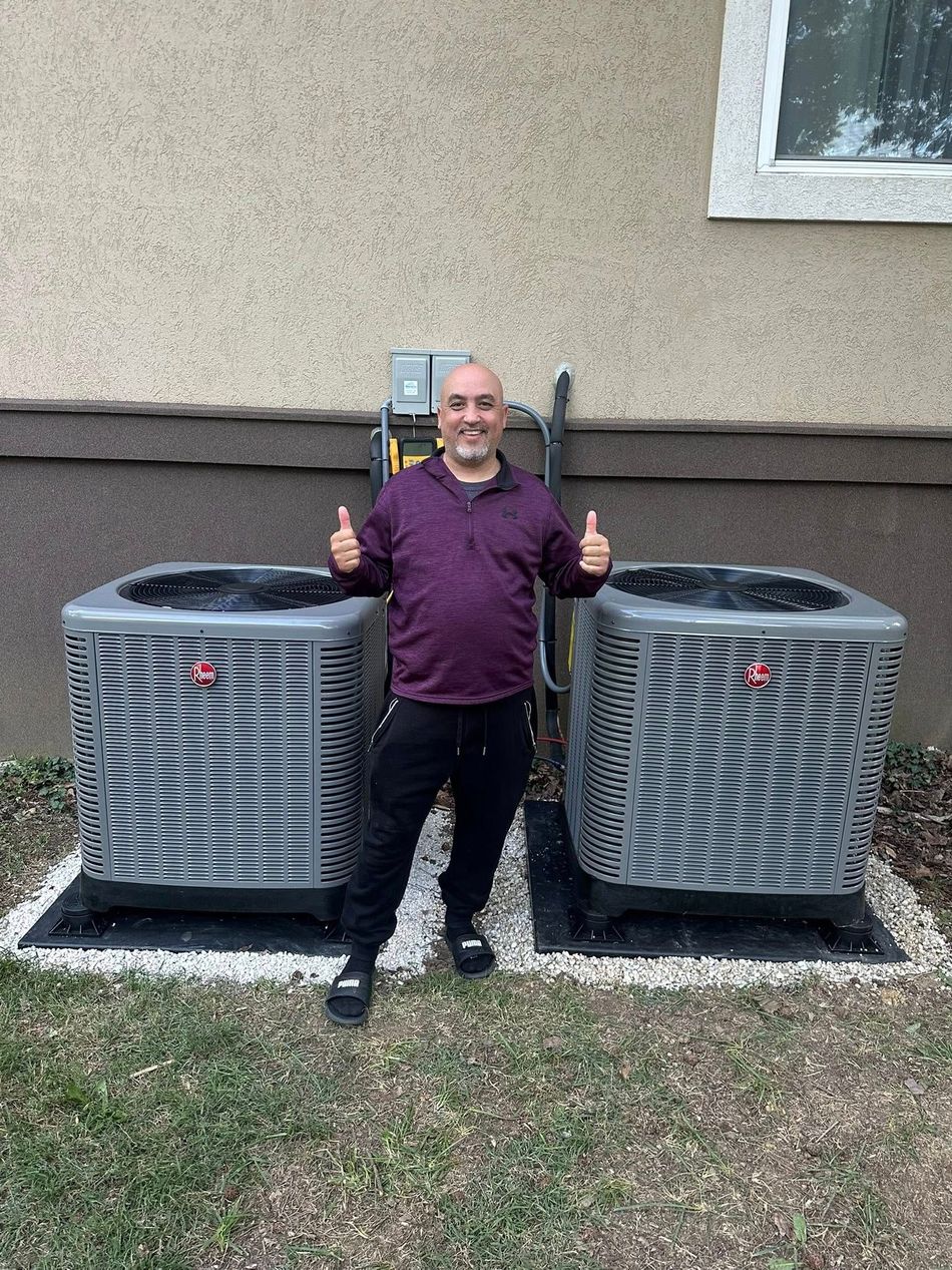Hudson’s Heating & Air Conditioning - A Man giving thumbs-up poses between two AC units outside a beige building.