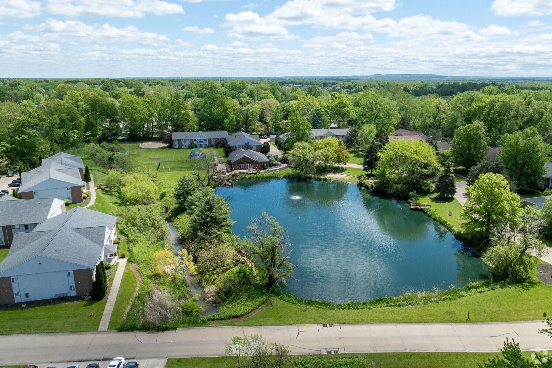 Aerial view of a pond surrounded by green trees, with houses in the background and a blue sky above.