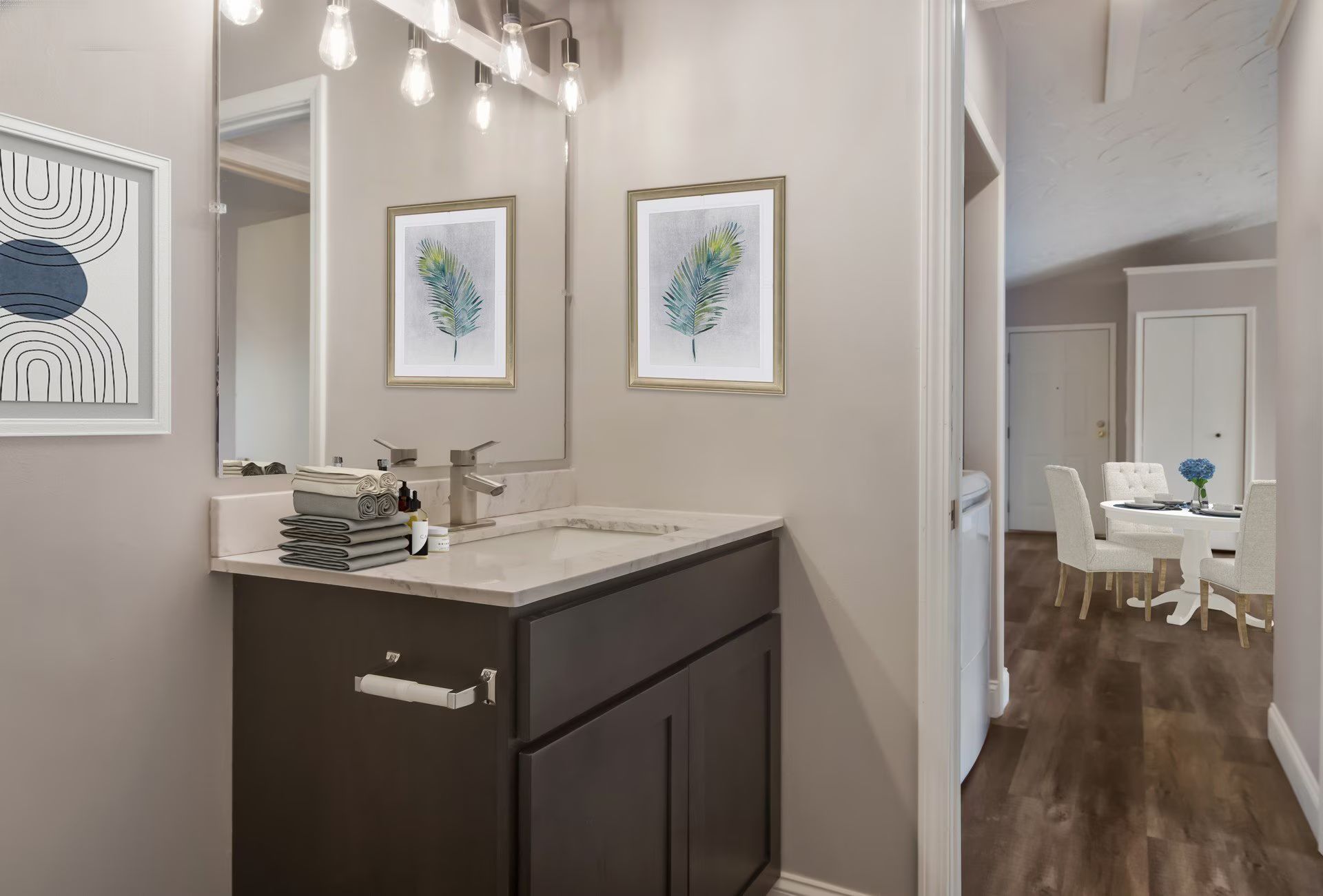 Bathroom with dark brown vanity, mirror, framed art, and doorway to dining area with table and chairs.