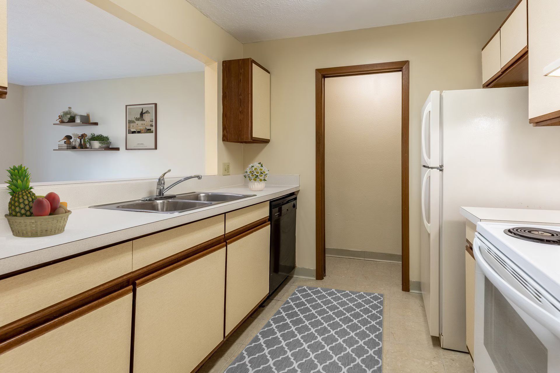 Kitchen with cream-colored cabinets, white appliances, and a gray patterned rug; a door leads to another room.