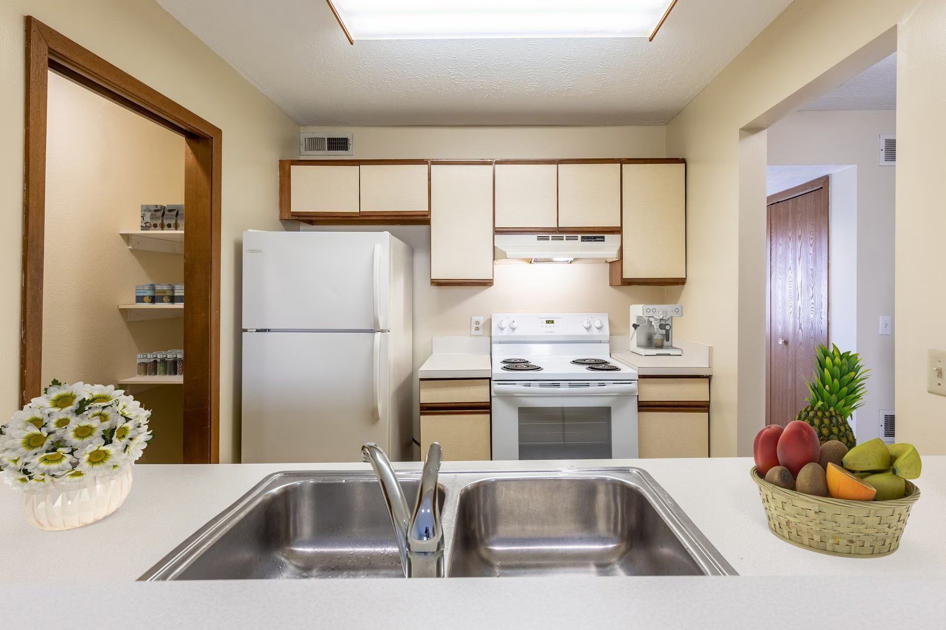 Kitchen with white appliances, light-colored cabinets, and a pantry to the left. A fruit basket and flowers are on the counter.