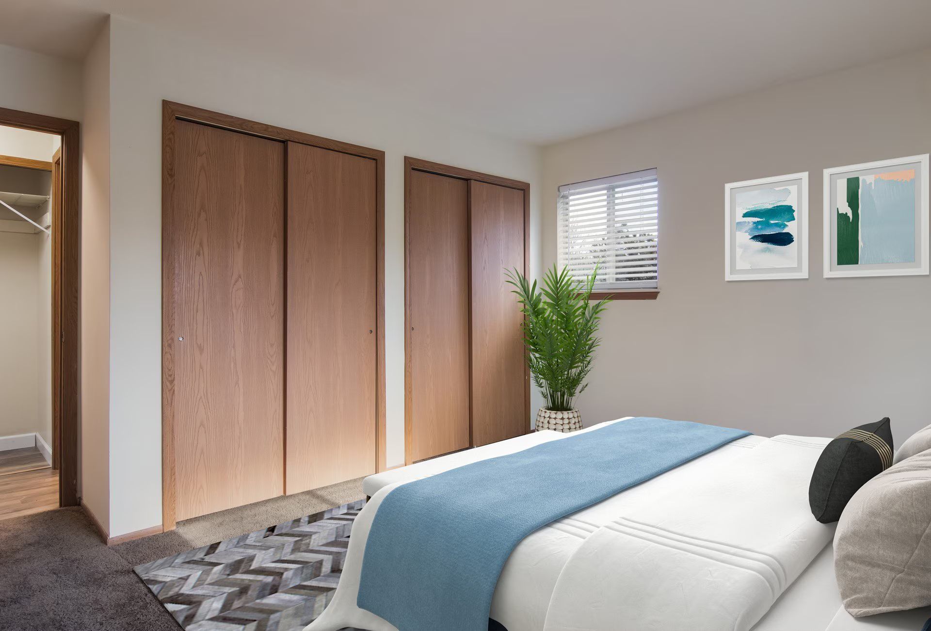 Bedroom with wood closet doors, bed with blue blanket, small window, art prints, and a potted plant.