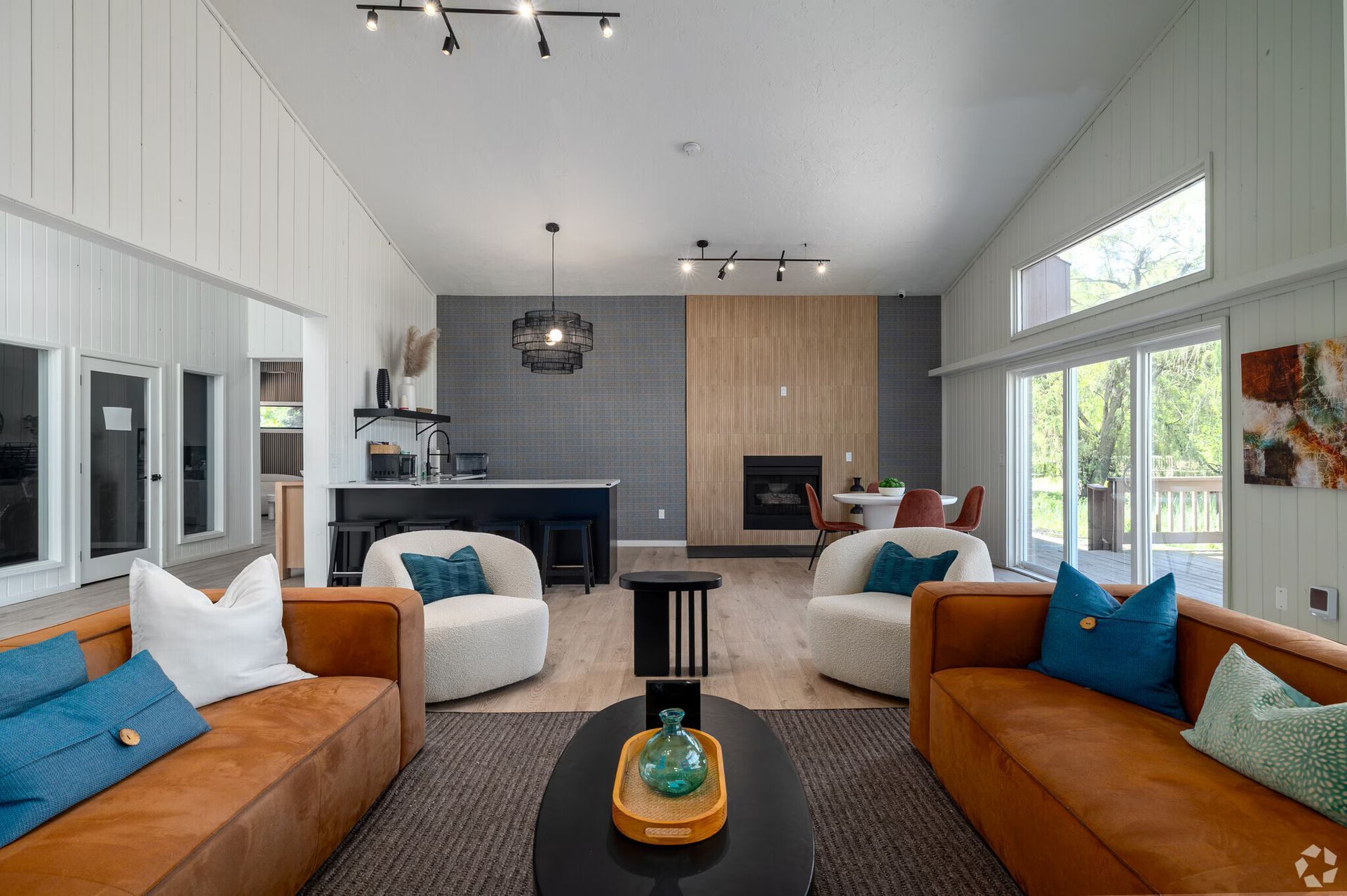 Open-plan living room with brown leather sofas, white armchairs, and a fireplace. Sliding glass doors lead to a deck.