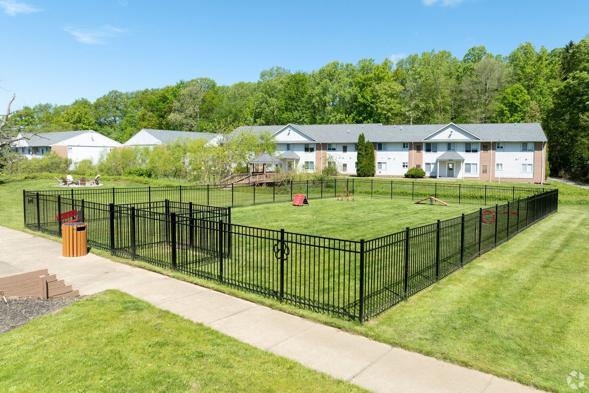 Fenced dog park with grassy area and apartment buildings in the background. Sunny day.