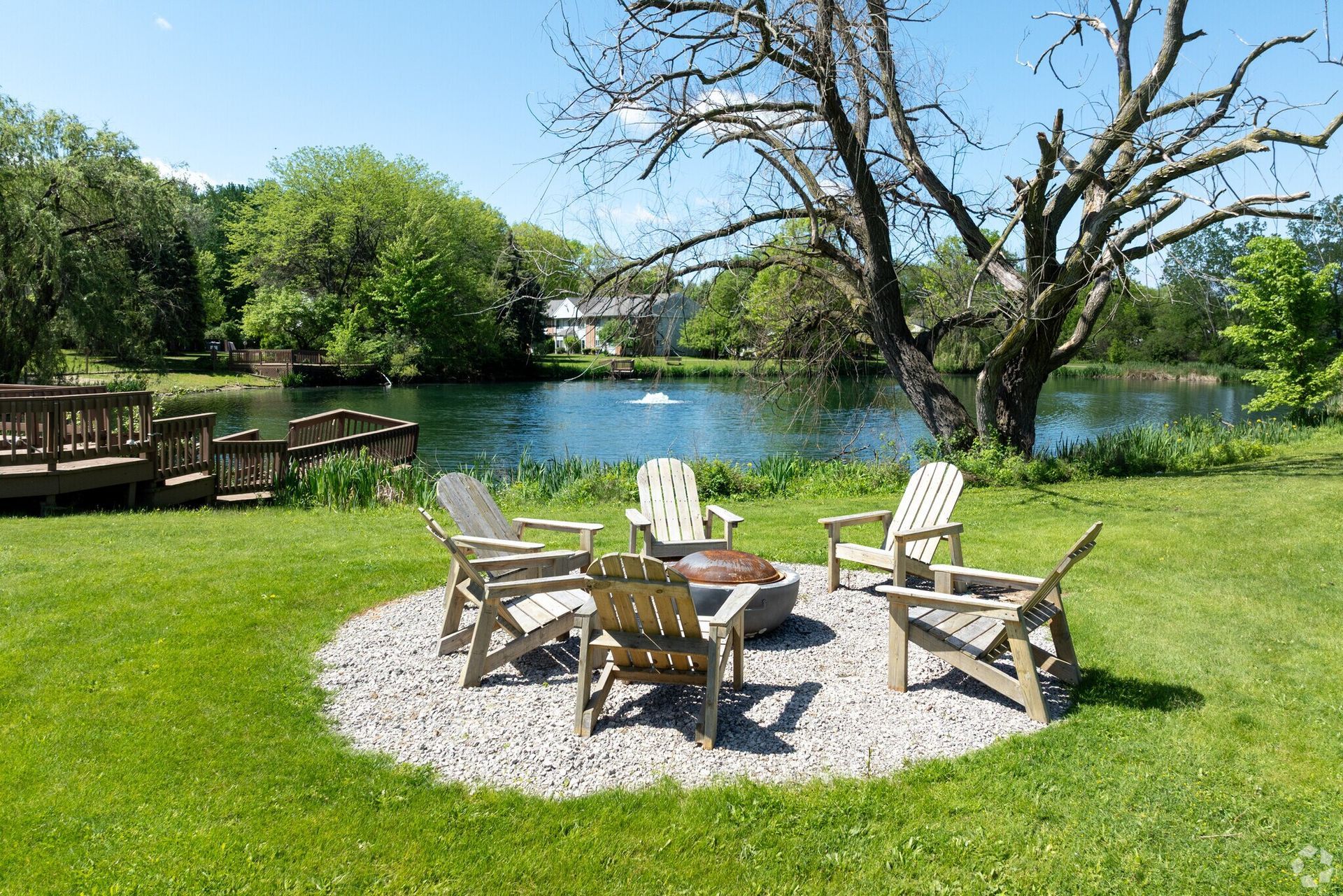 Six wooden chairs around a fire pit on a gravel base, overlooking a pond.