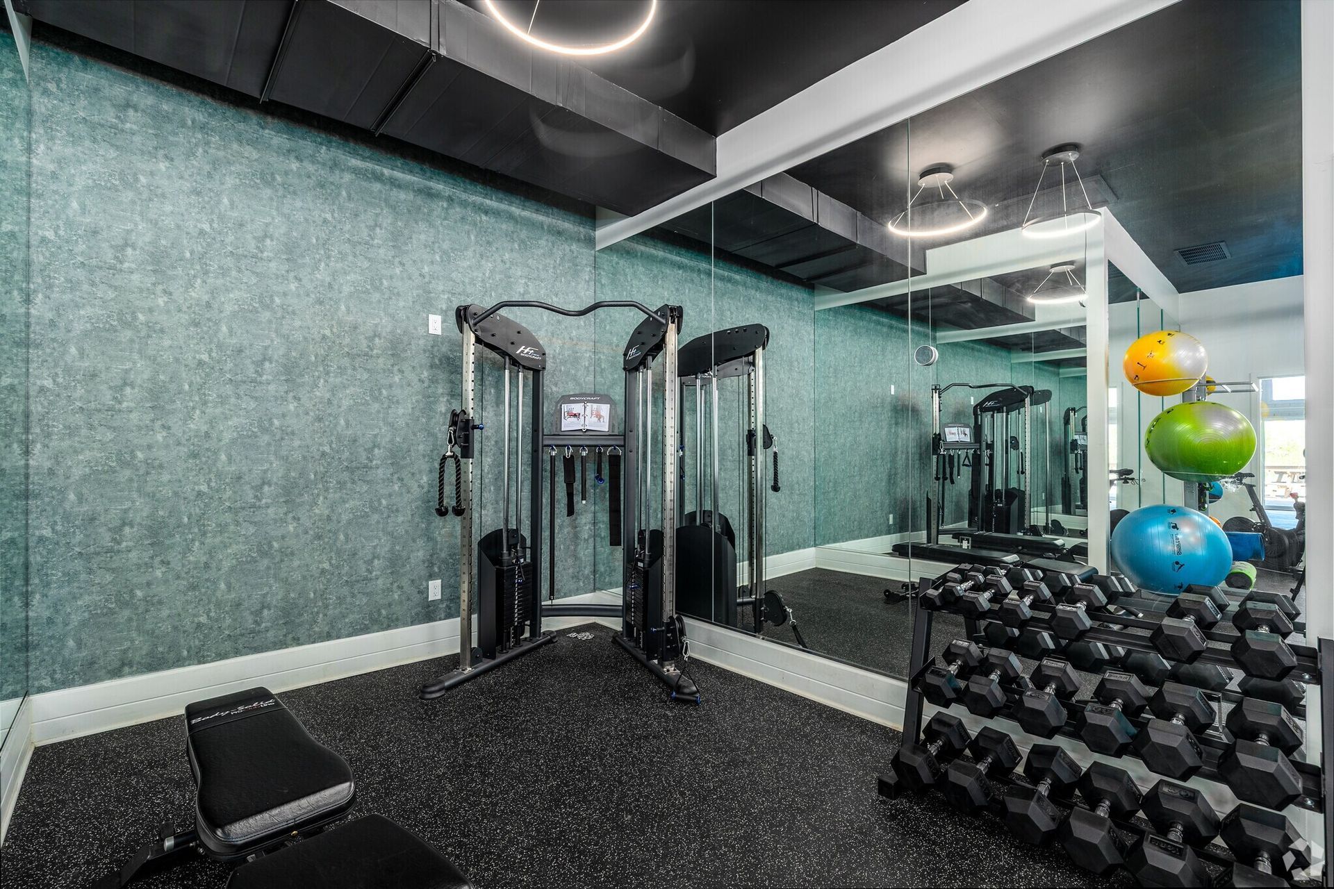 Gym with weight machine, dumbbells, bench, and exercise balls. Mirrored wall reflects equipment.