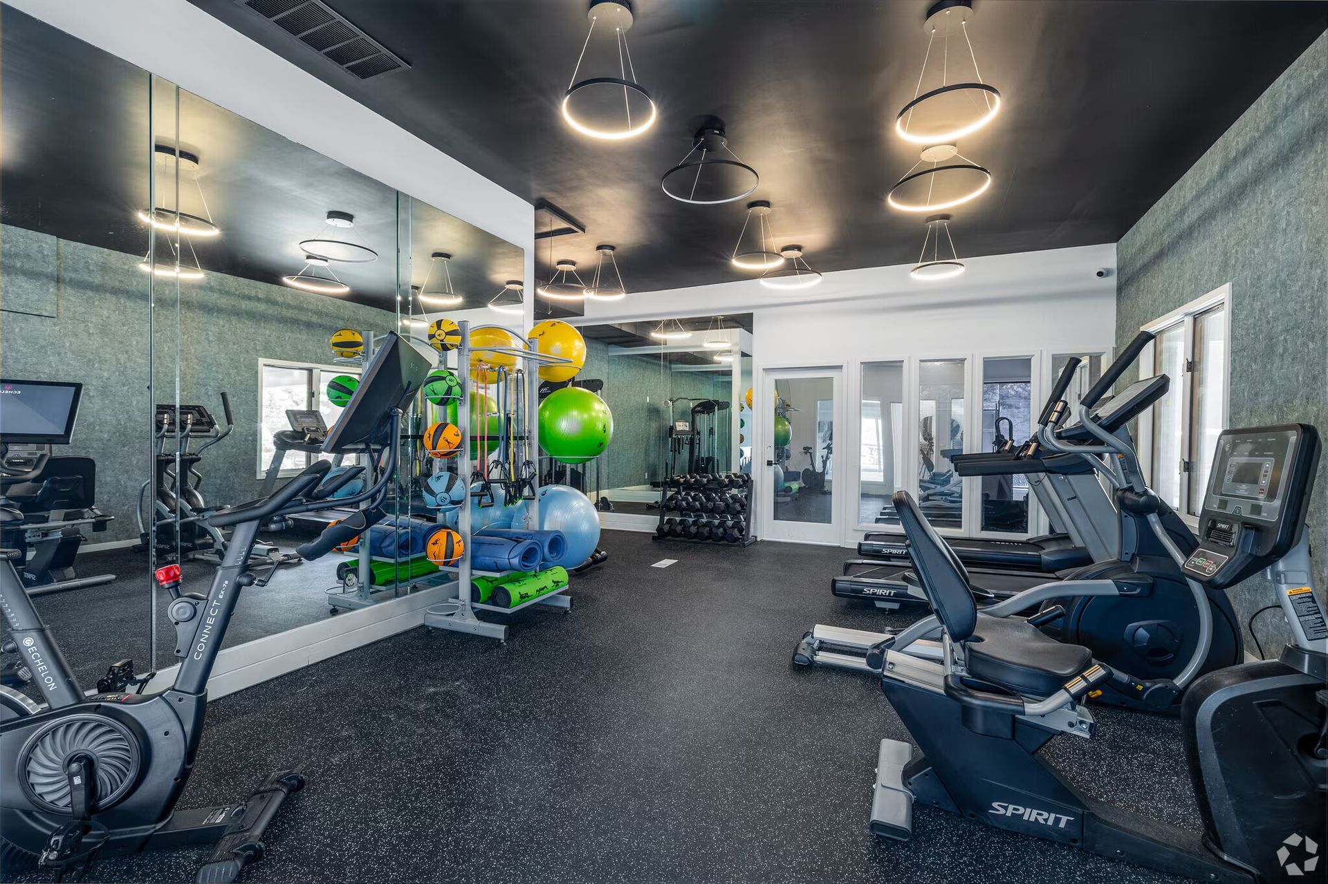 Gym with exercise equipment: stationary bikes, treadmills, free weights, and fitness balls. Mirrors and overhead lights.