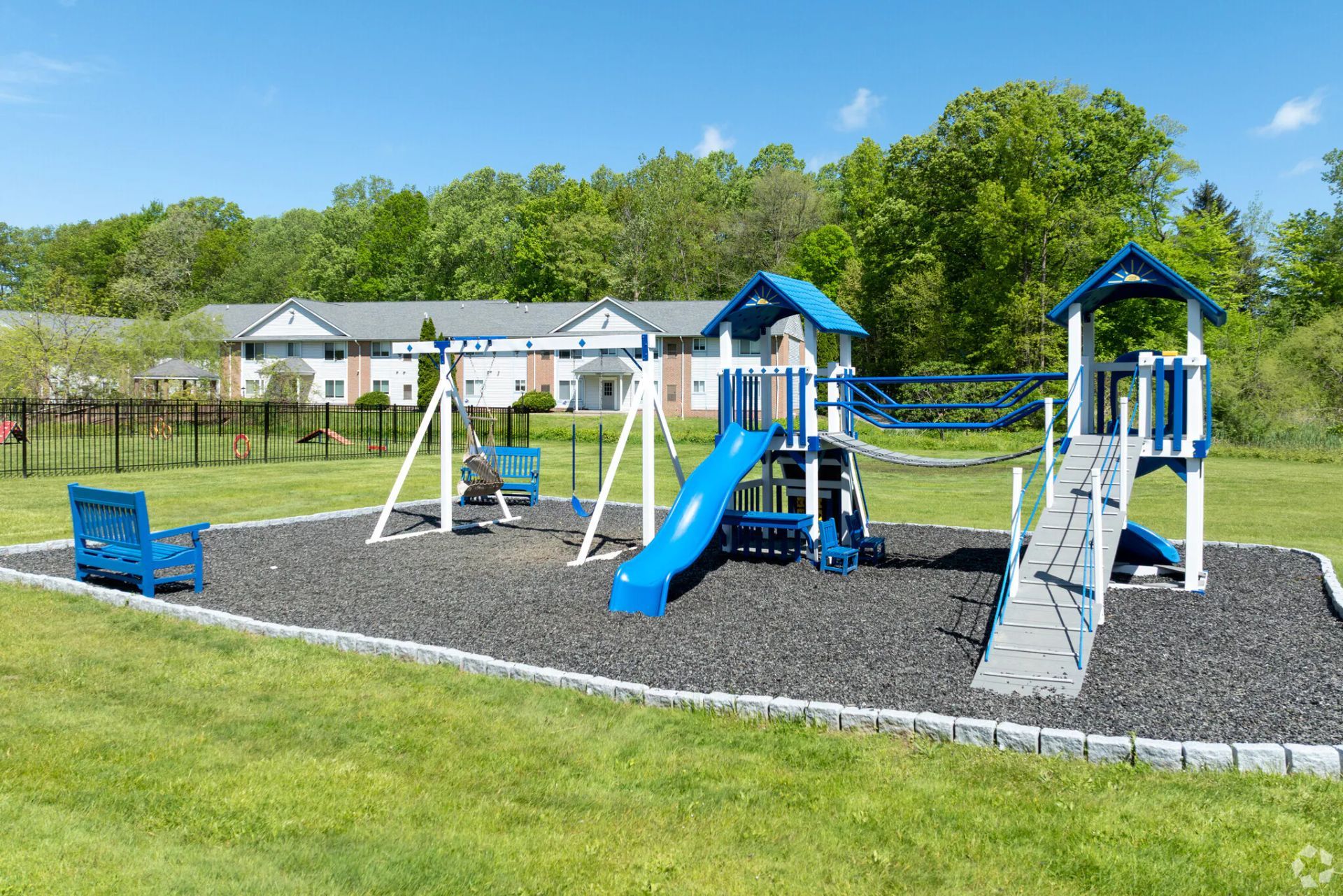 Playground with blue and white equipment on a black rubber surface. Green lawn, trees, and buildings in the background.
