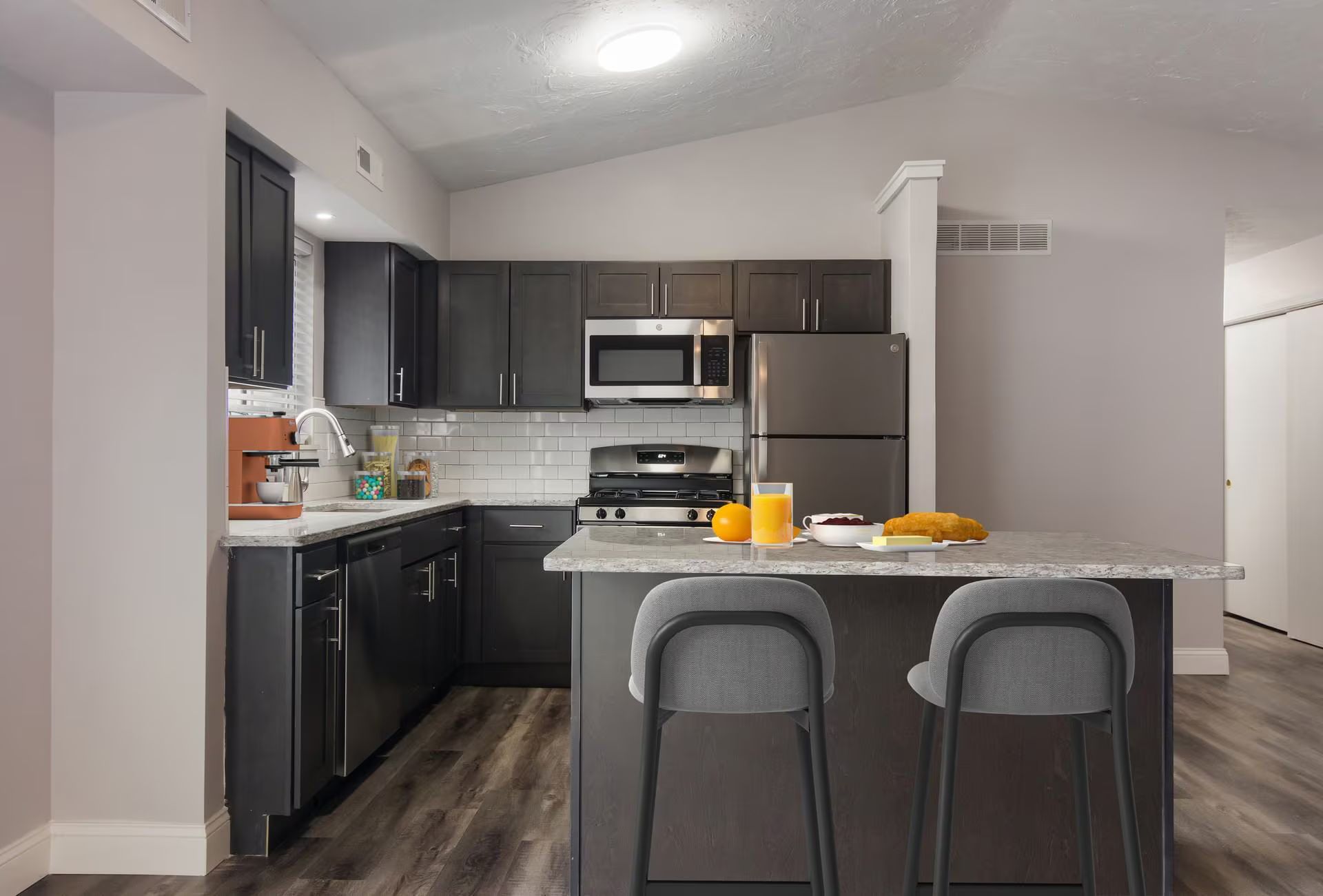 Modern kitchen with dark cabinets, stainless appliances, and a breakfast bar with two gray stools.