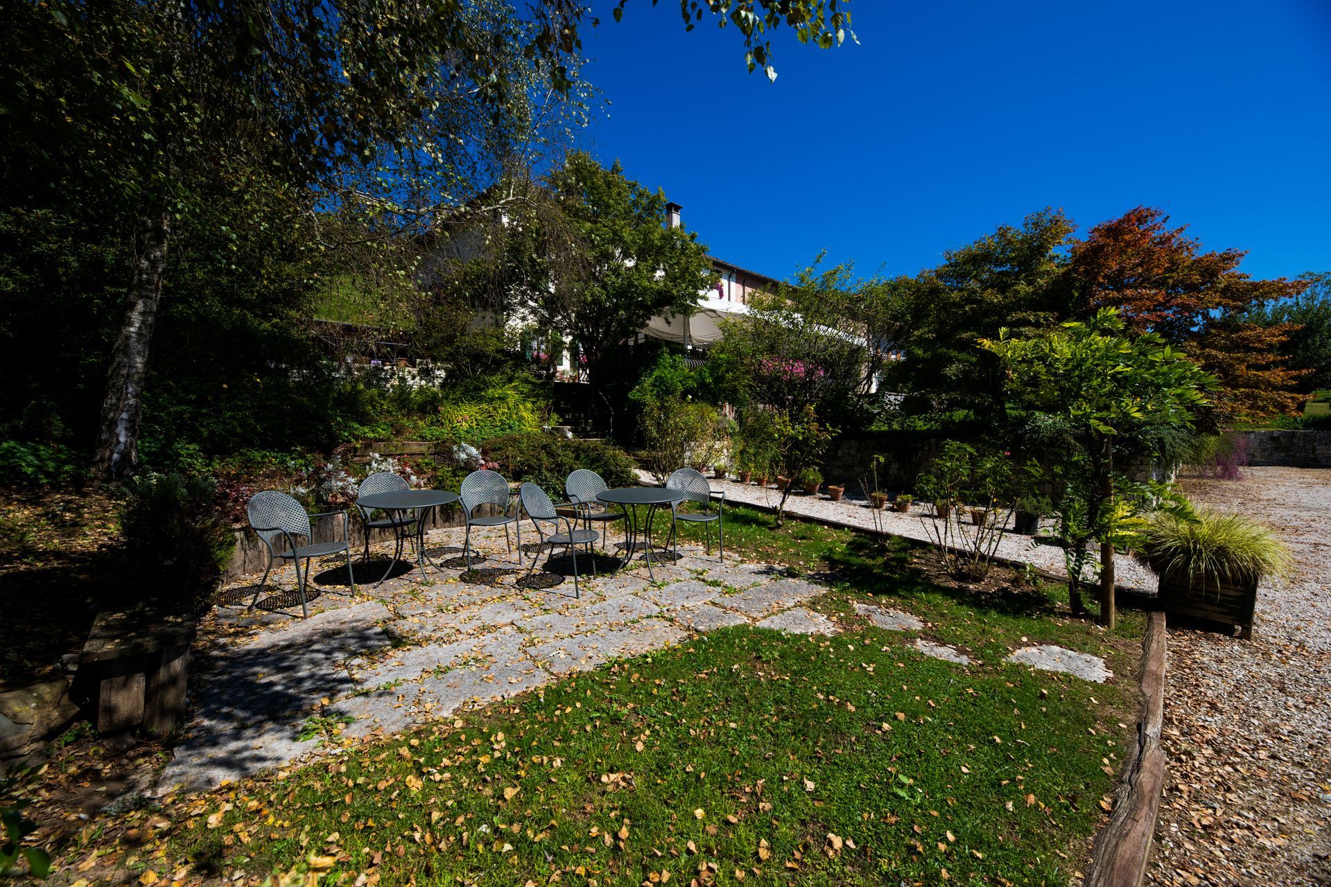 Zona pranzo all'aperto con tavoli e sedie, alberi ed edificio nelle giornate di sole.