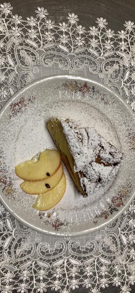 Fetta di torta su un piatto con fette di mela, spolverata di zucchero a velo. Piatto su una tovaglia decorativa.
