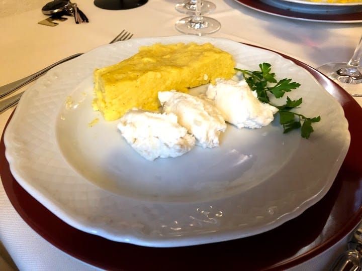 Polenta con formaggio e prezzemolo su un piatto bianco, adagiata su un piatto rosso, con posate d'argento.