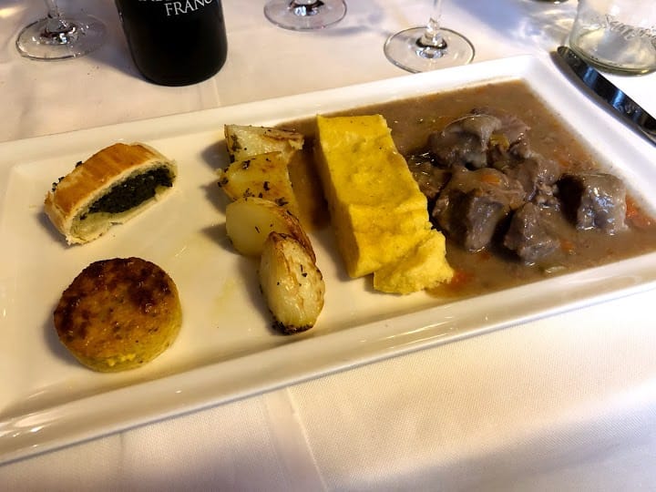 Piatto di cibo con carne in salsa, polenta, patate e rotolo di pasta sfoglia su una tovaglia bianca.