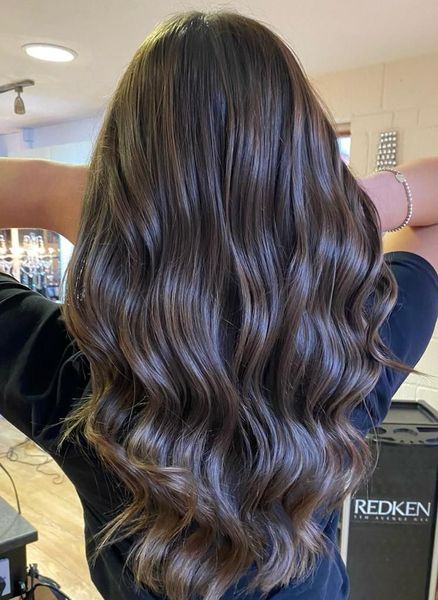 A woman with long brown hair is standing in a salon with her arms outstretched.