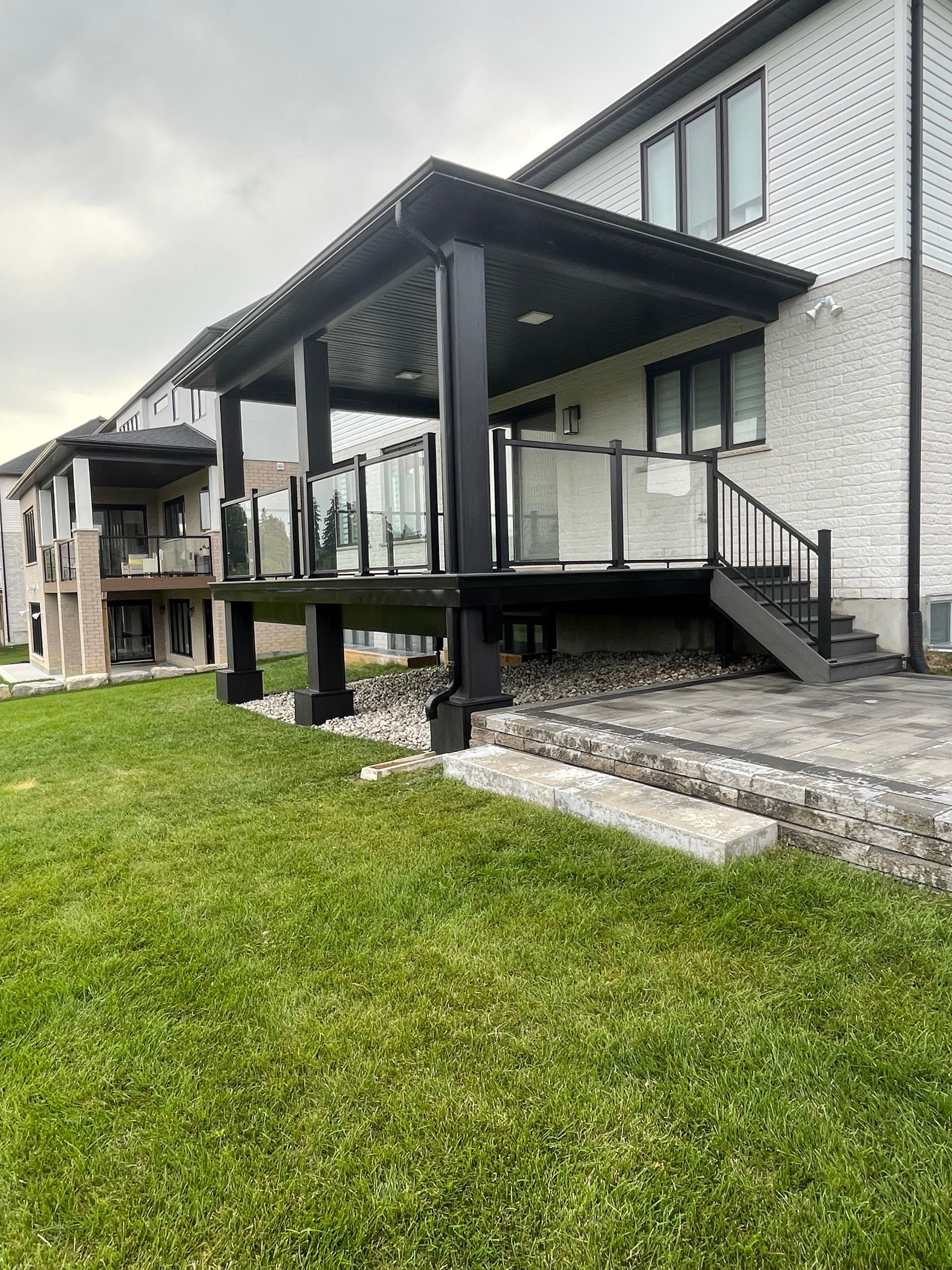 Black deck with glass railings and overhead roof, attached to a two-story brick house, gray sky.