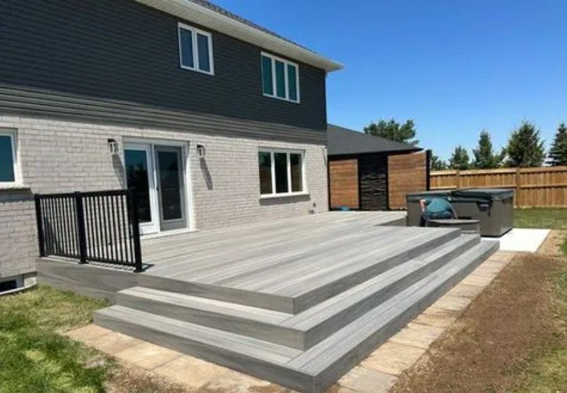 Gray composite deck with steps, black railing, hot tub, against a gray brick house on a sunny day.