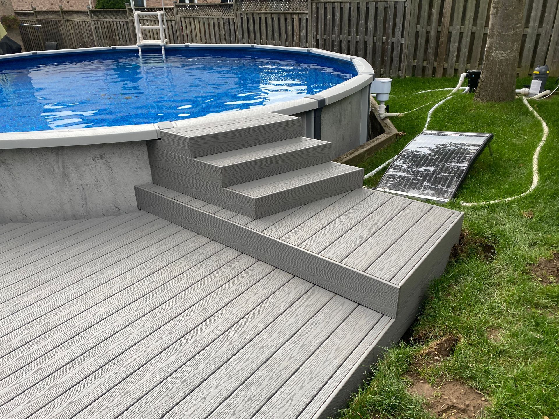 Above-ground pool with attached grey composite deck and steps. A solar panel sits nearby on the grass.