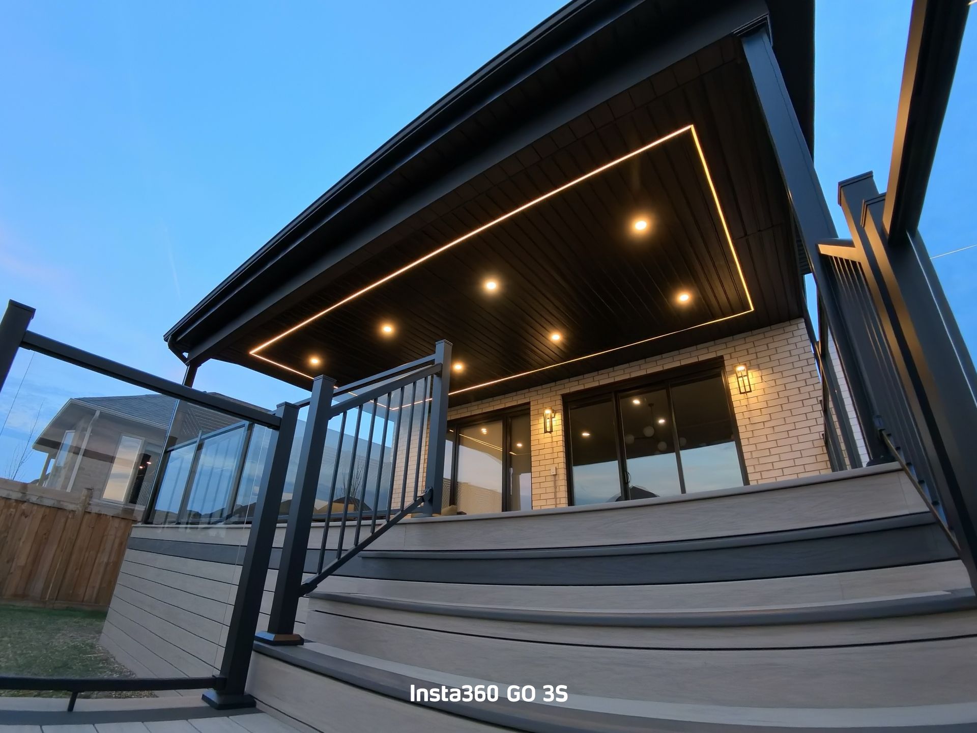 Exterior view of a deck with stairs, black railing, and a covered ceiling with lights; evening.