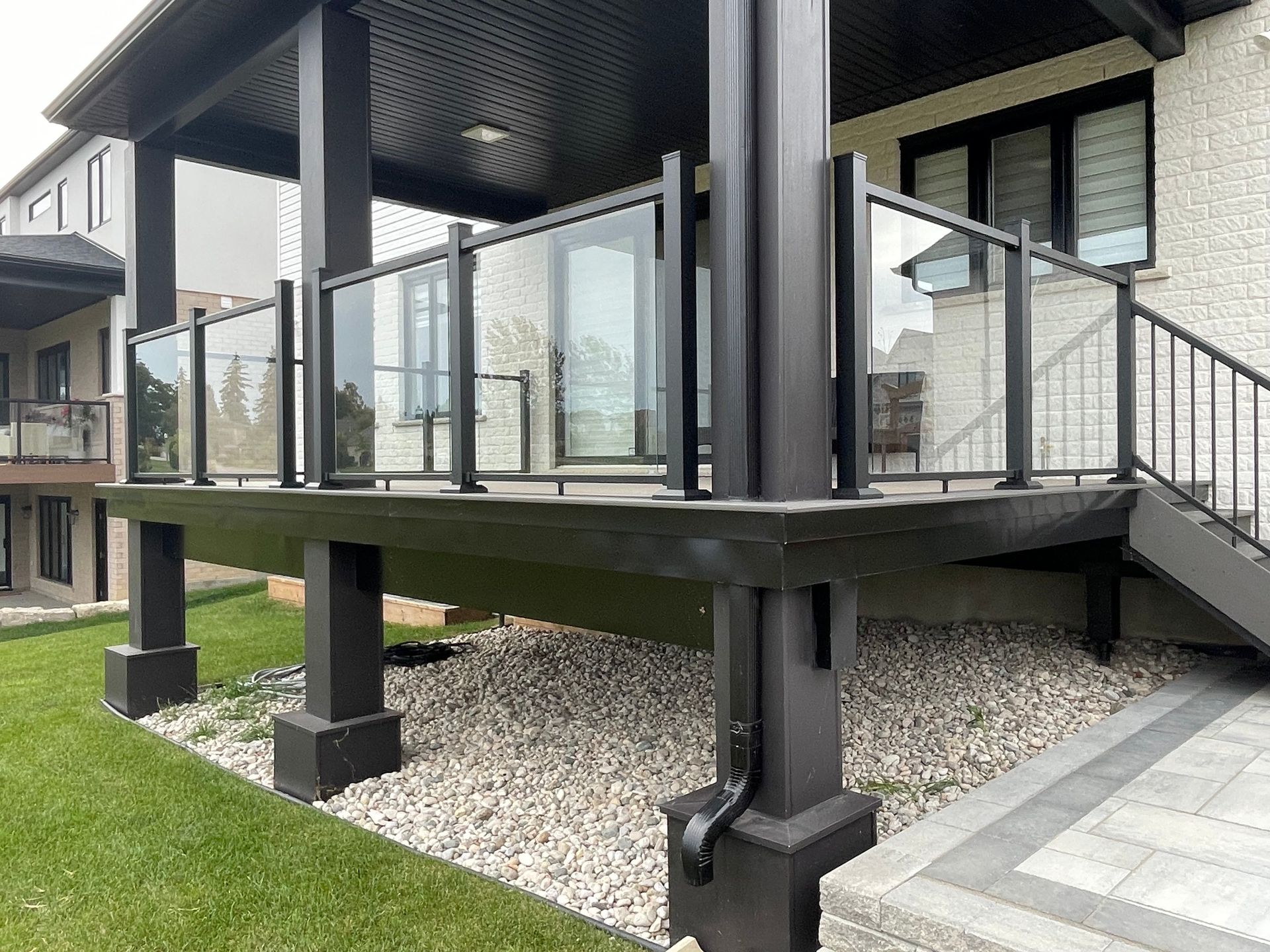 Black deck with glass railings, supported by dark columns, next to a stone patio and grassy lawn.