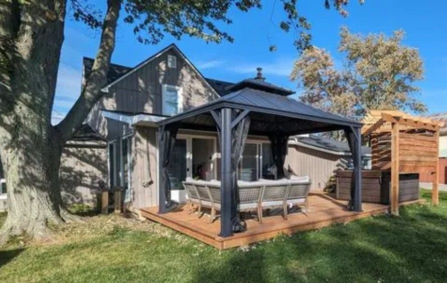 Backyard patio with gazebo, deck, and house under a blue sky and tree.