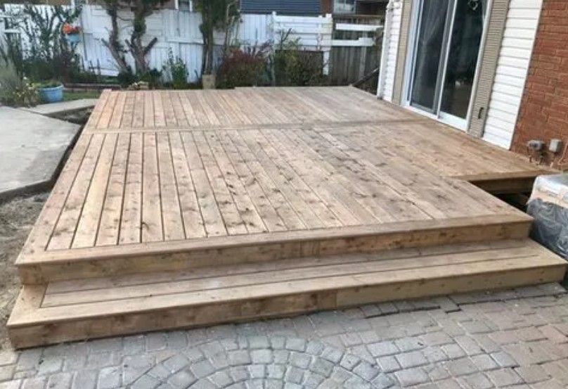 Wooden deck with steps leading to a paved patio, beside a brick house and sliding glass door.