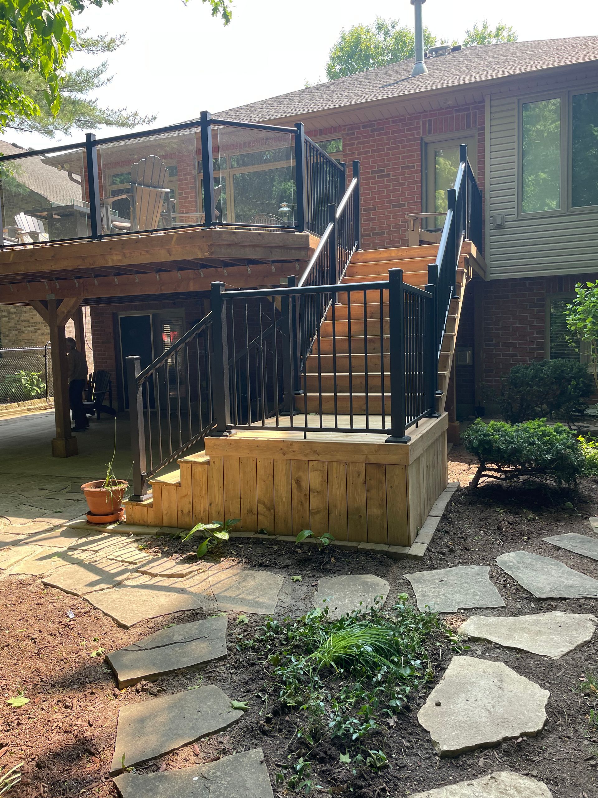 Wooden deck with stairs, black railing, glass panels, attached to a brick house. Landscaping in foreground.