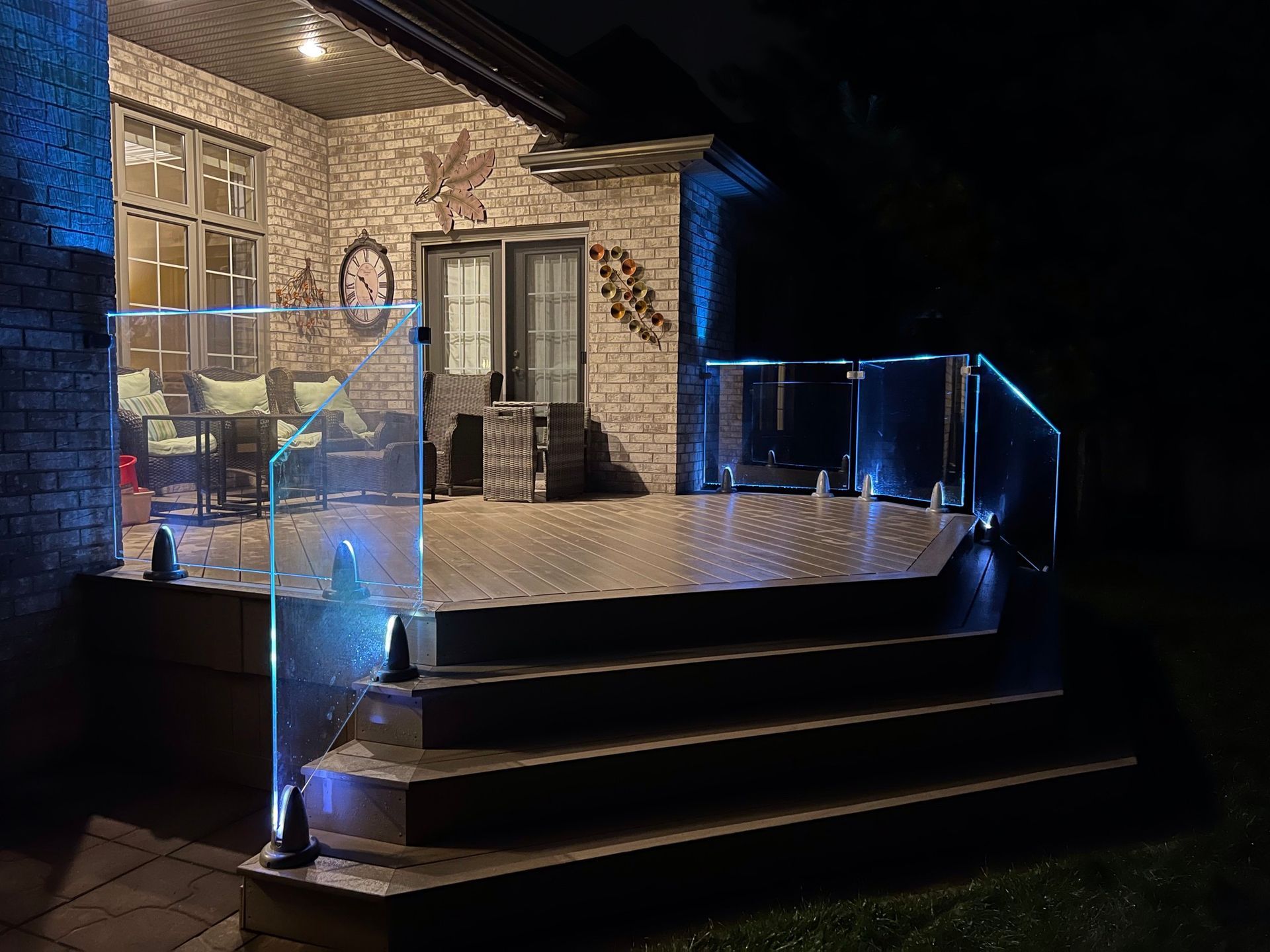 Lit deck at night with blue lights on glass railing and steps. Brick house, lounge furniture visible.
