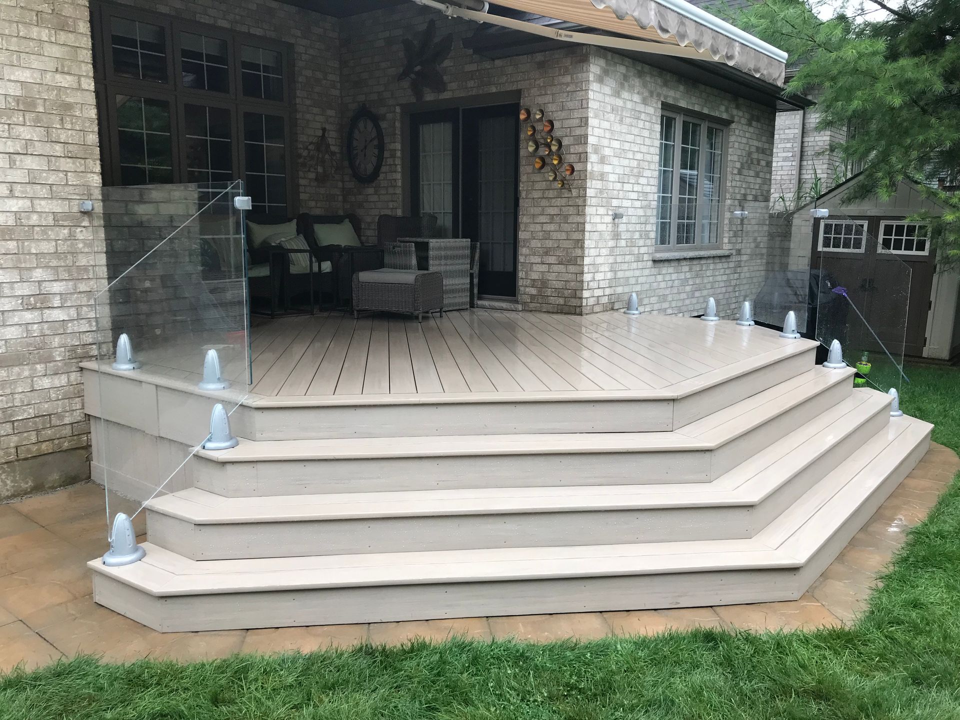 Outdoor deck with multiple steps, glass railing, light brown composite decking, and brick house.
