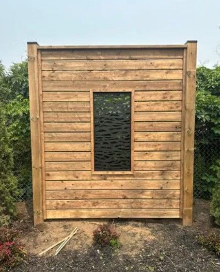 Wooden privacy screen with rectangular window; set in a garden with plants and overcast sky.