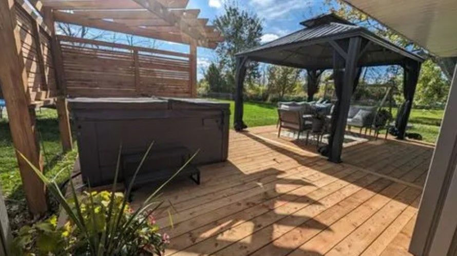 Outdoor wooden deck with hot tub and gazebo. Sunny day, green grass and trees in background.