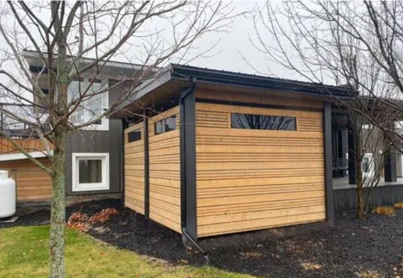 Wooden shed-like structure attached to a house with horizontal wood panels, black trim, small windows, and a black roof.