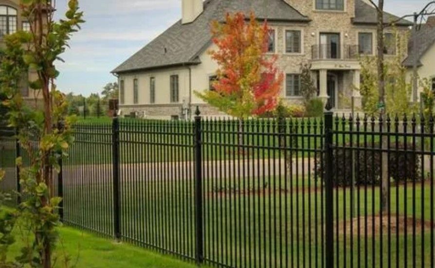 Black metal fence surrounding a large house with a green lawn and trees in fall.