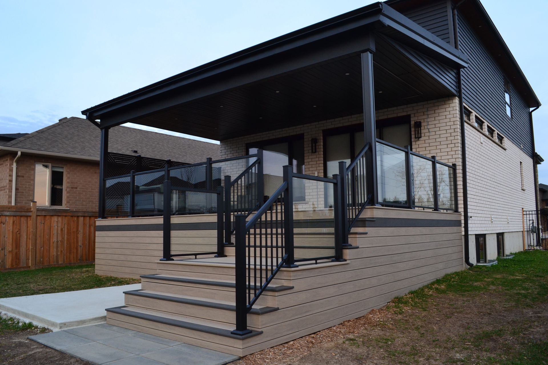 House with deck and glass railings; steps lead to covered porch, siding in neutral tones.