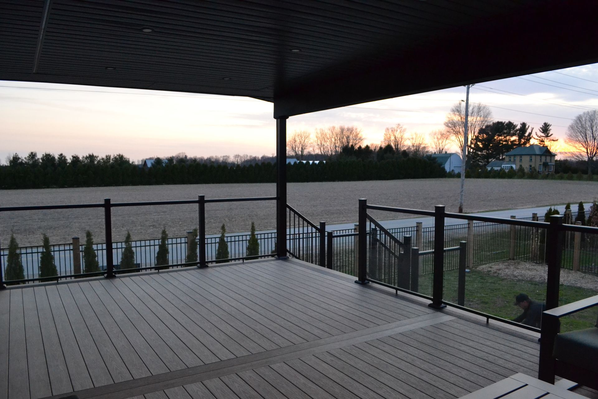 A wooden deck with black railings overlooks an open field, trees, and a house at sunset.
