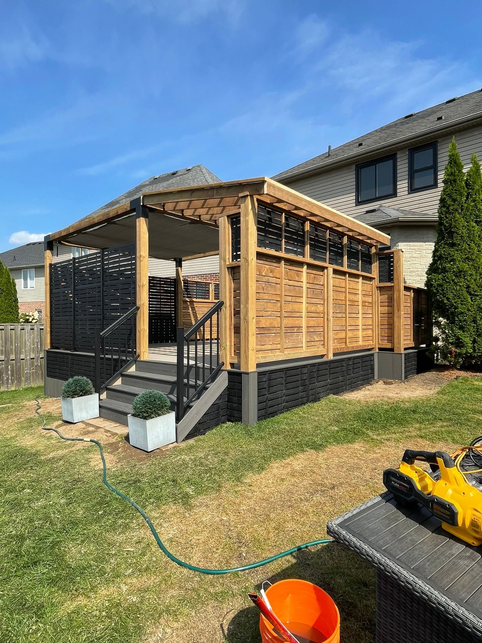 Backyard deck with wooden pergola and lattice, attached to a two-story house. Green lawn, blue sky.